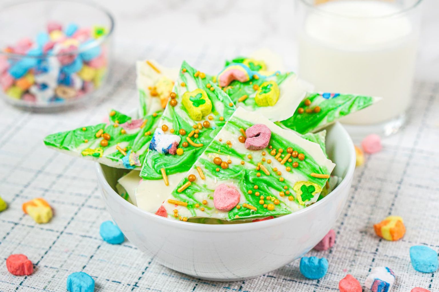 A white bowl filled with pieces of white and green candy bark topped with colorful cereal and sprinkles, with a glass of milk and a bowl of cereal pieces in the background.