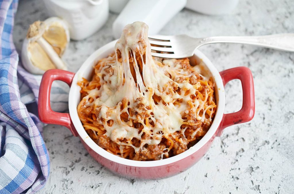 A red baking dish filled with baked pasta topped with melted cheese, with a fork lifting a cheesy portion. Garlic and a blue-checked cloth are nearby on a light countertop.