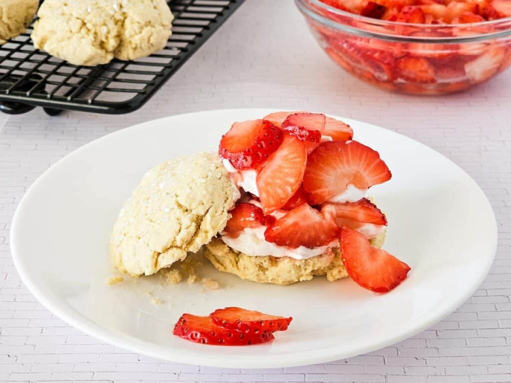 A biscuit split open and topped with whipped cream and sliced strawberries on a white plate, with more biscuits and strawberries in the background.