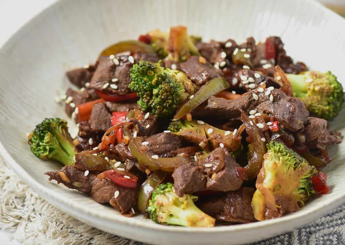 A plate of beef stir-fry with broccoli, bell peppers, onions, and sesame seeds on a light-colored dish.