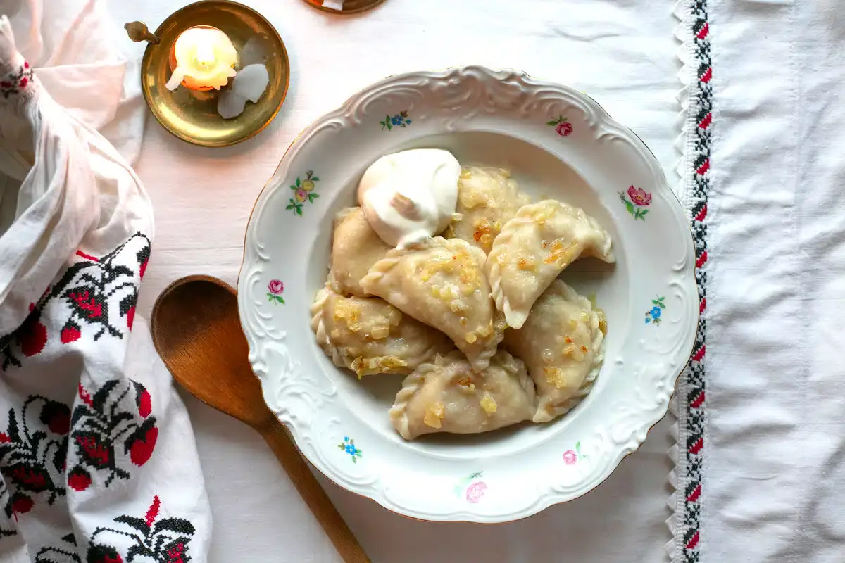 A plate of dumplings topped with fried onions and a dollop of sour cream, beside a wooden spoon, candle, and embroidered cloth on a white tablecloth.