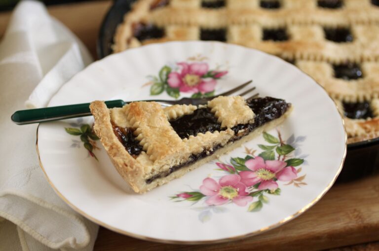 A slice of lattice-topped fruit pie on a floral plate with a fork, next to the remaining pie in the background.