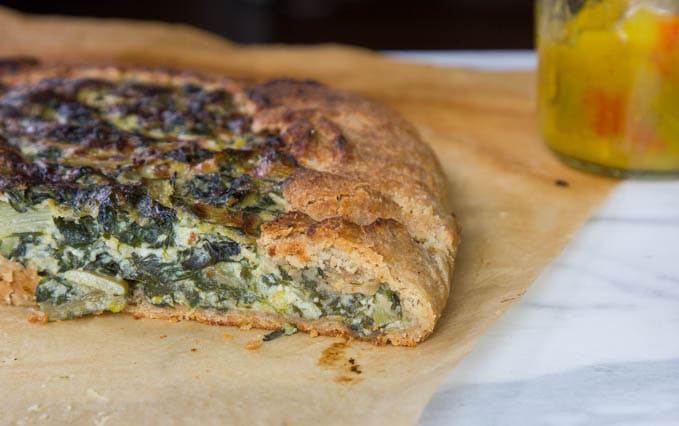 A partially sliced savory galette with a vegetable and cheese filling sits on parchment paper next to a glass jar containing pickled vegetables.