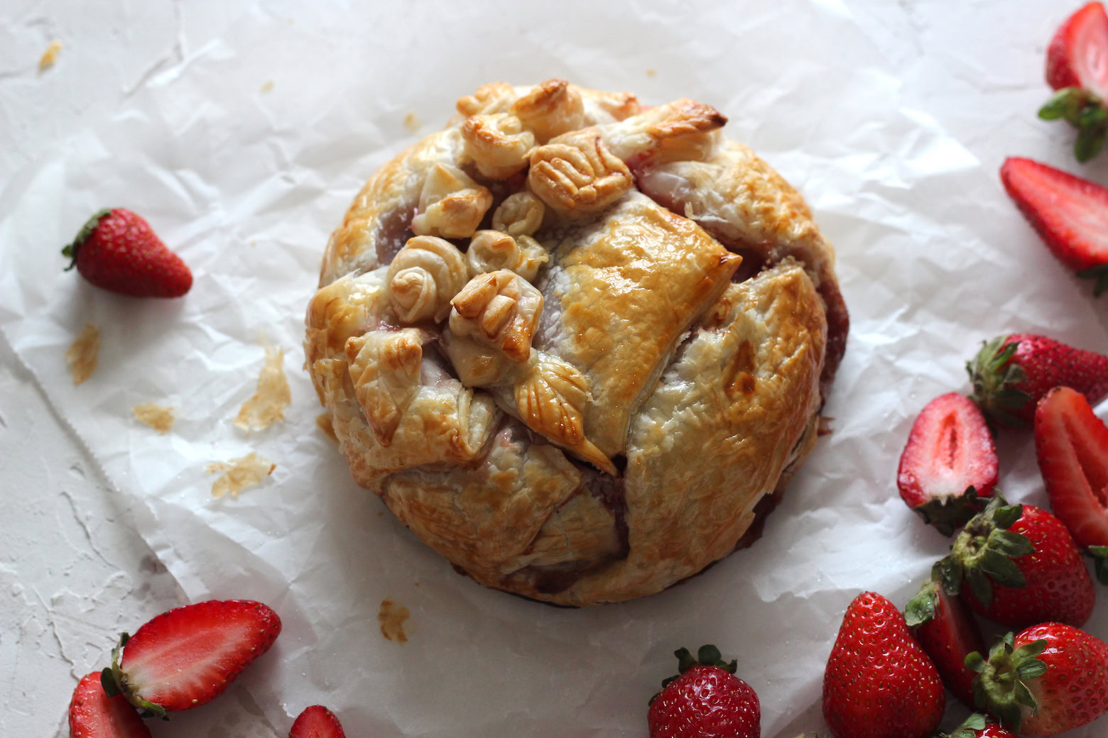 A round baked pastry with decorative shapes on top is placed on parchment paper, surrounded by whole and halved strawberries.