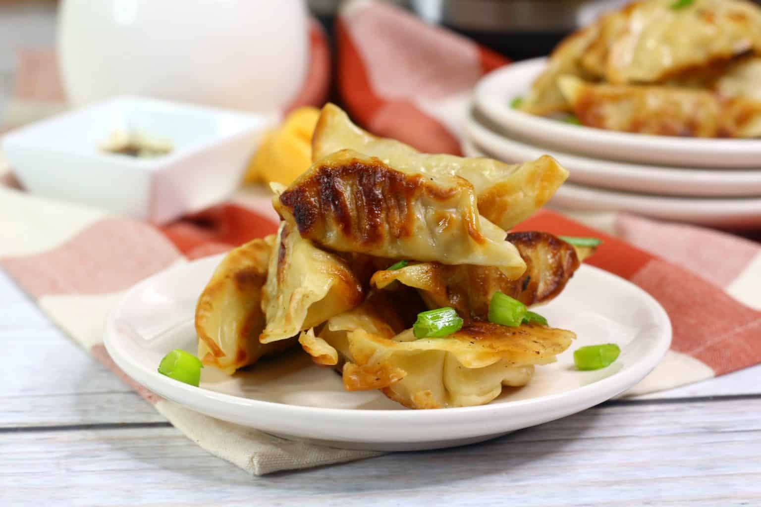 A small plate of pan-fried dumplings garnished with chopped green onions, with more dumplings and dipping sauce in the background.