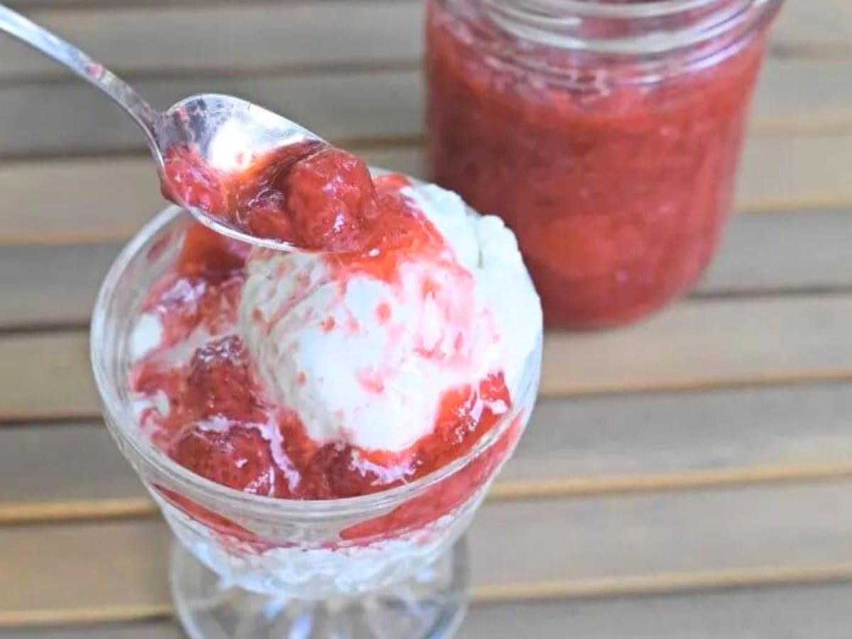 A spoonful of strawberry compote is being placed on vanilla ice cream in a glass dish, with a jar of compote in the background.