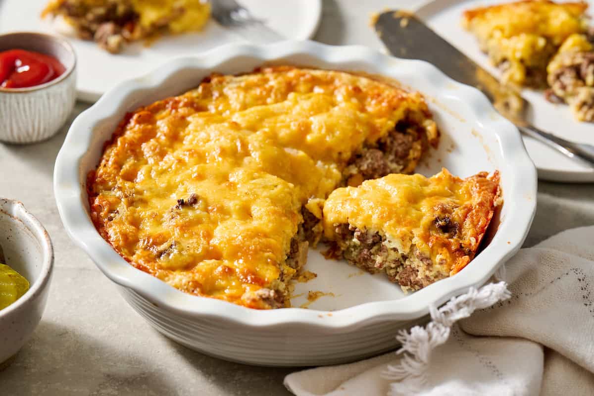 A baked casserole with a golden, melted cheese topping, partially sliced, in a white dish on a light surface with plates and condiments nearby.