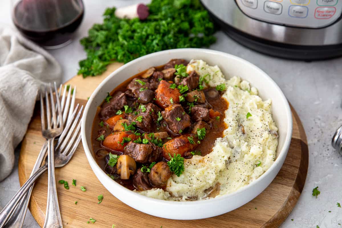 A bowl of beef stew with carrots, mushrooms, and gravy served alongside mashed potatoes, garnished with chopped parsley, placed on a round wooden board with forks nearby.