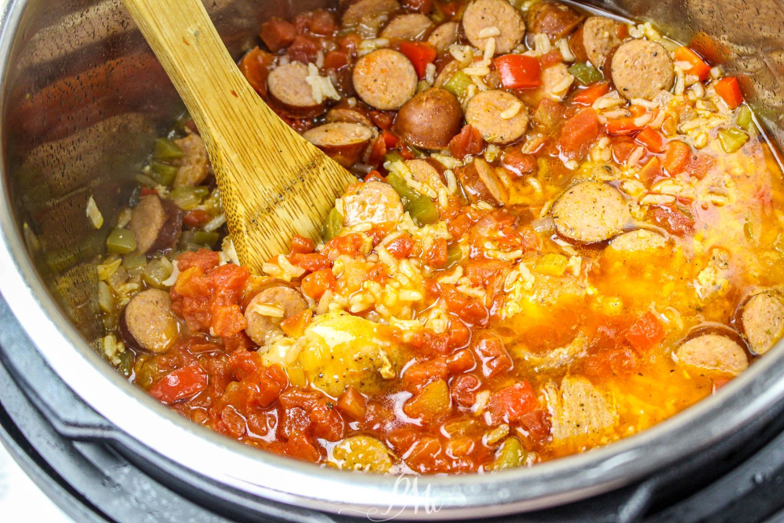 A wooden spoon stirs a mixture of sliced sausage, tomatoes, rice, celery, and peppers inside an Instant Pot.