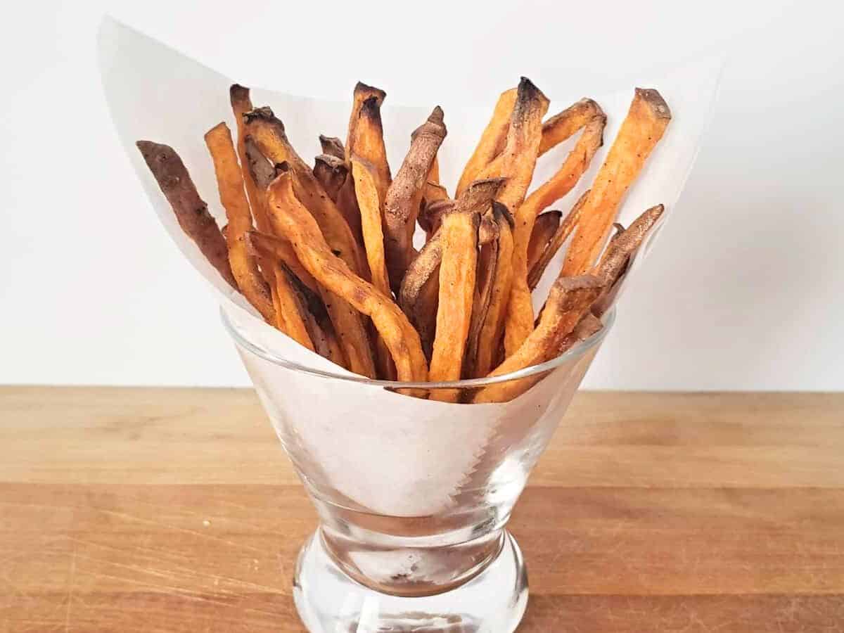 Sweet potato fries arranged upright in a parchment-lined glass on a wooden surface with a plain white background.