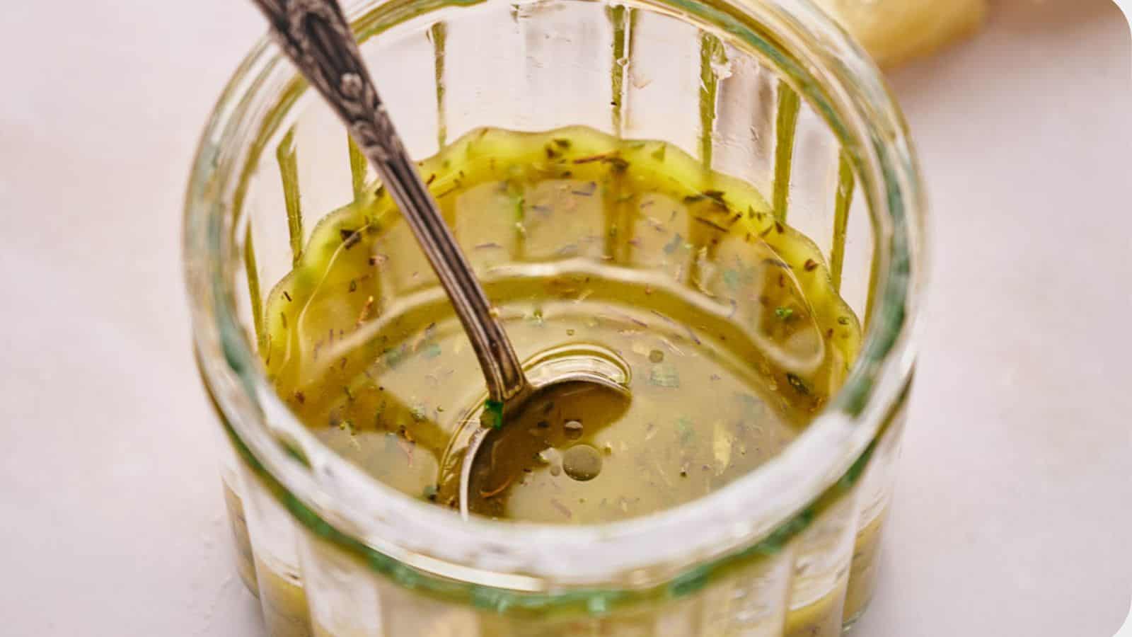 A glass jar filled with a yellow-green herb vinaigrette, with a metal spoon resting inside.