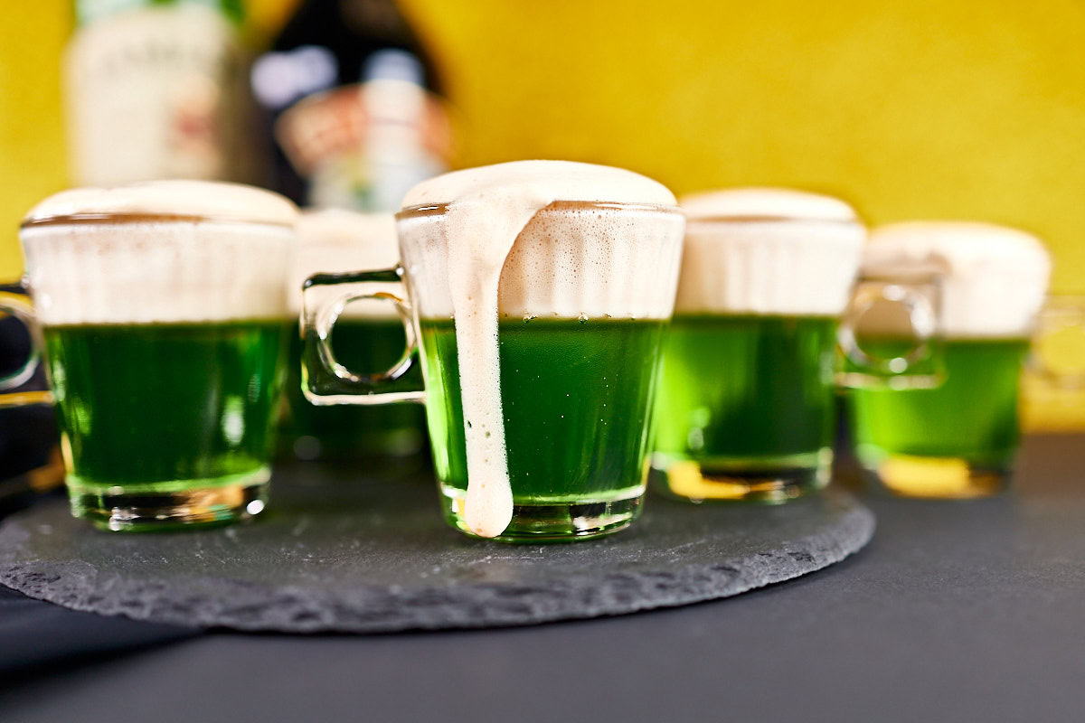 Several glass mugs filled with green liquid topped with frothy foam, set on a dark surface with a yellow background; one mug has foam spilling over the side.
