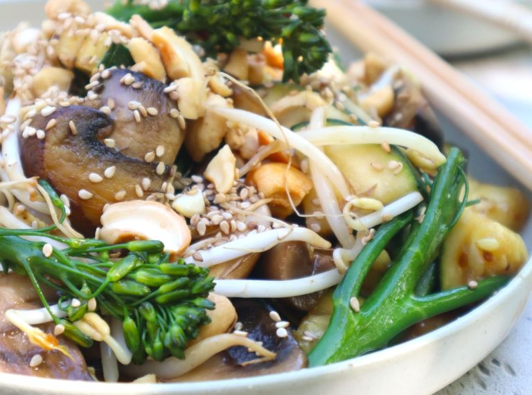 A close-up of a vegetable stir-fry with mushrooms, broccolini, bean sprouts, cashews, and sesame seeds, served on a plate.