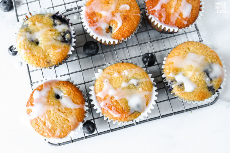 Six blueberry muffins with white icing sit on a wire cooling rack, with a few loose blueberries nearby.