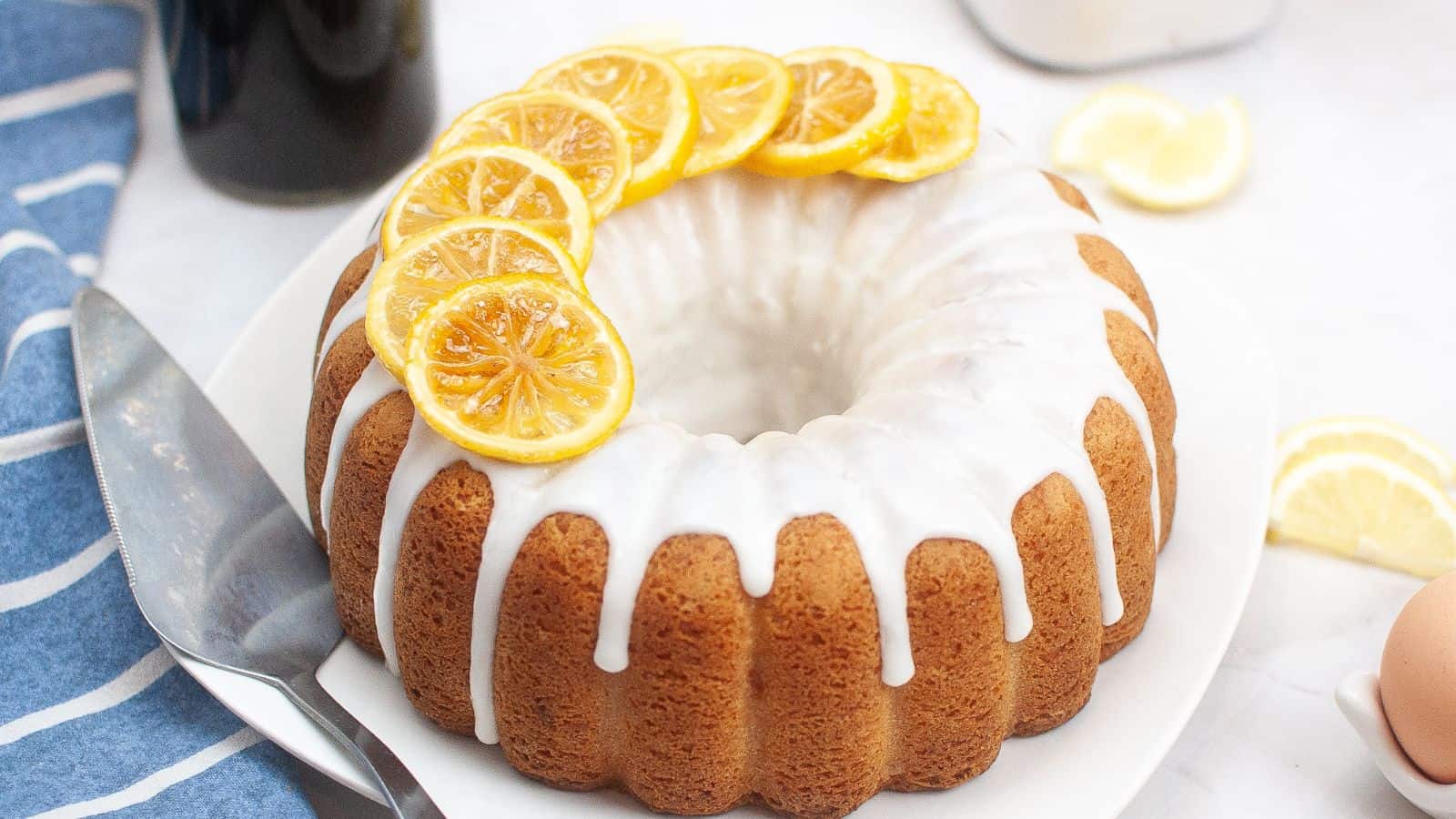 A bundt cake with white icing drizzled on top, garnished with sliced lemons, sits on a white plate next to a cake server and a blue-striped cloth.