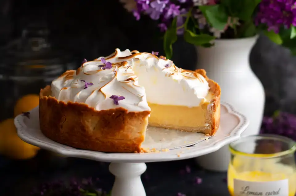 A lemon meringue pie with a slice removed sits on a cake stand, topped with browned meringue and purple flower petals; flowers and a jar of lemon curd are in the background.