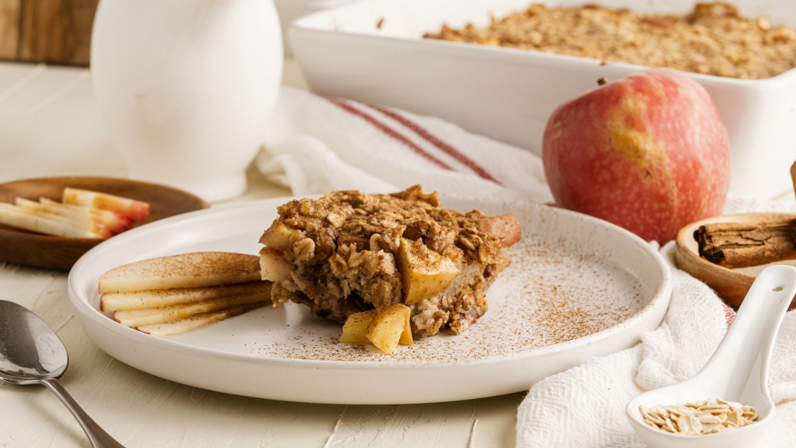 A slice of apple crumble sits on a white plate next to apple slices, with a whole apple and a baking dish in the background.