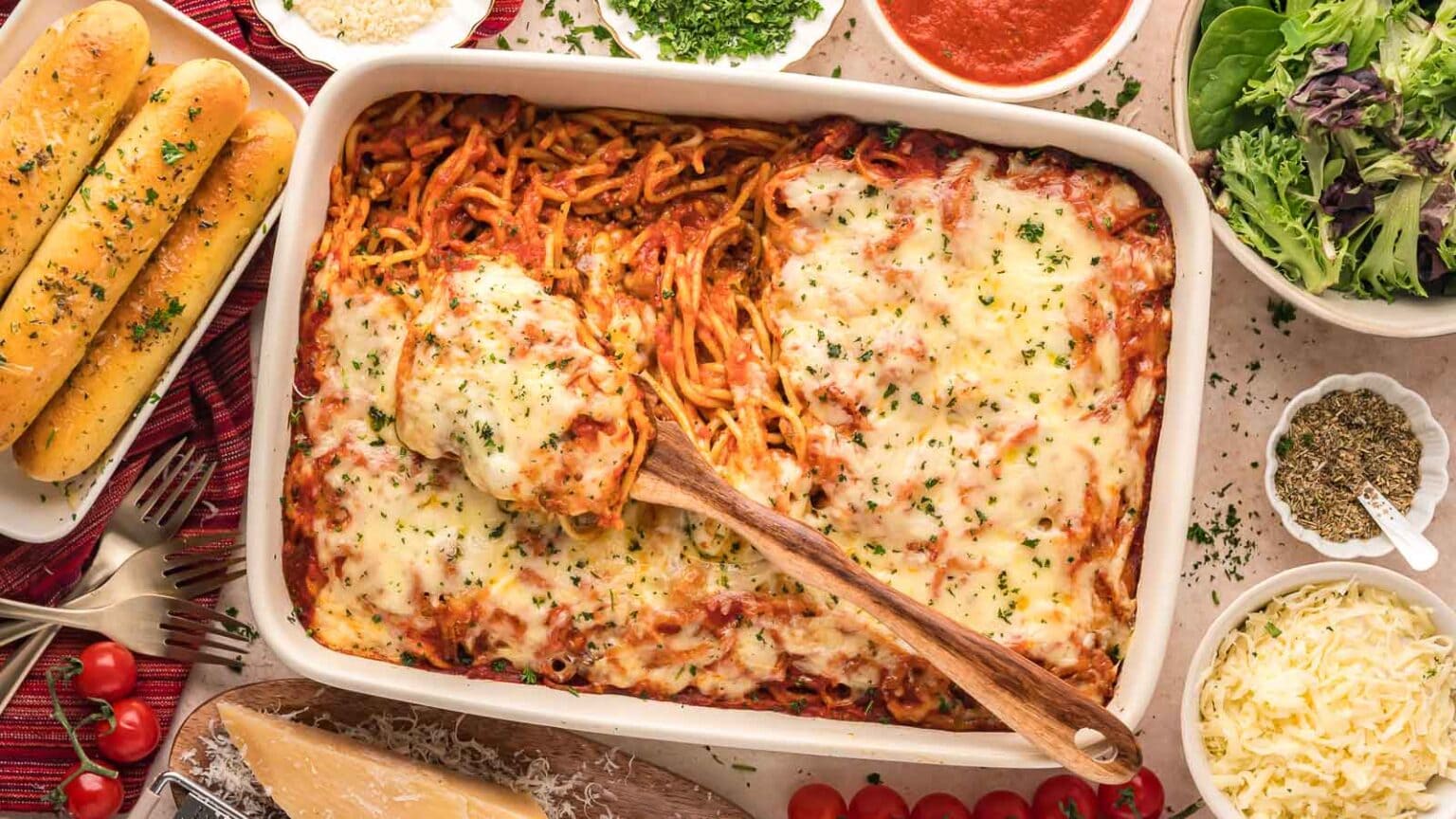 A baked spaghetti casserole with melted cheese is served in a white dish, surrounded by breadsticks, salad, grated cheese, herbs, and tomato sauce.