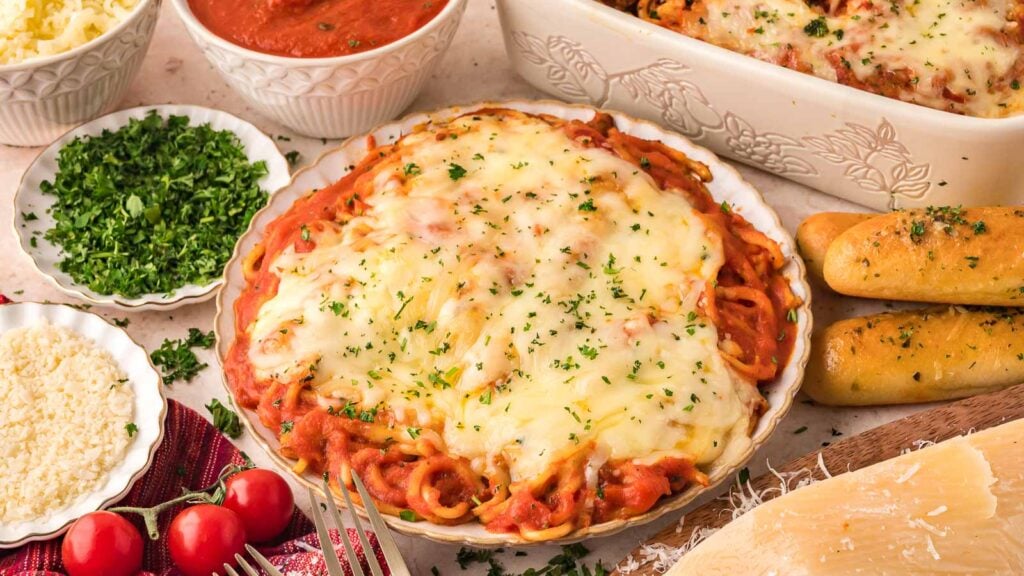 A plate of baked spaghetti topped with melted cheese, surrounded by breadsticks, chopped herbs, grated cheese, tomato sauce, and a baking dish of pasta.
