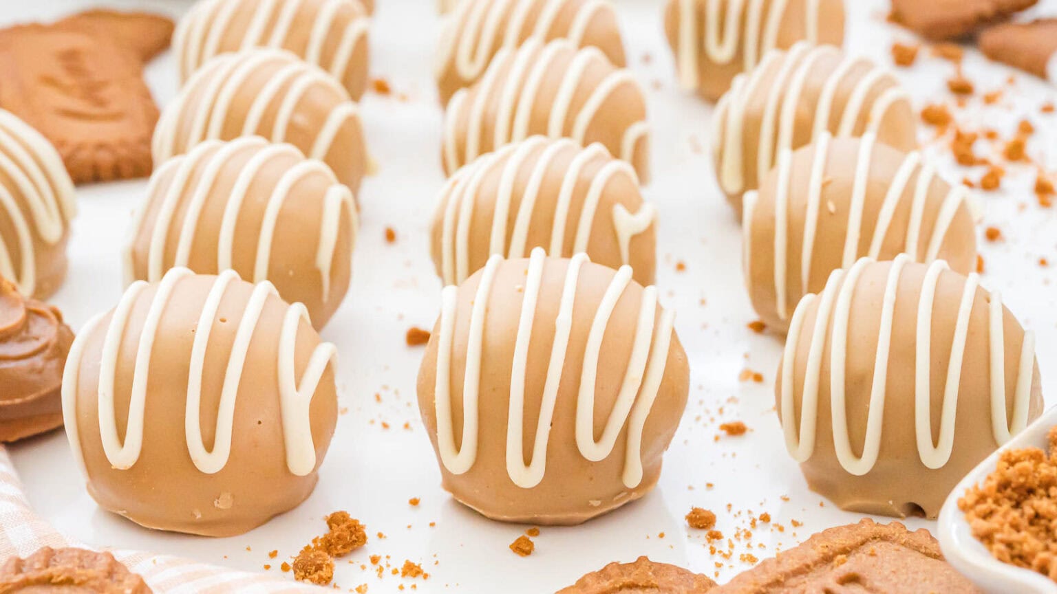 Rows of round, tan-colored truffles drizzled with white icing, arranged on a white surface with cookie crumbs and pieces scattered around.