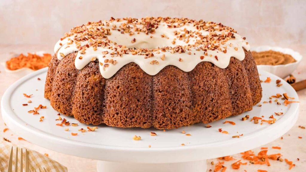 A round carrot bundt cake with white icing and chopped nuts on top, displayed on a white cake stand.