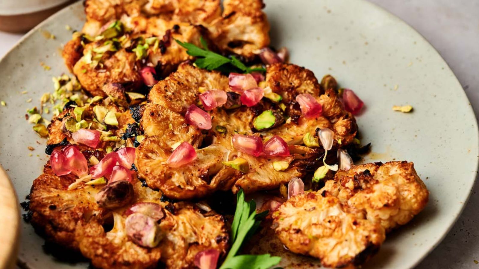 Roasted cauliflower steaks topped with pomegranate seeds, chopped pistachios, and fresh herbs on a light-colored plate.