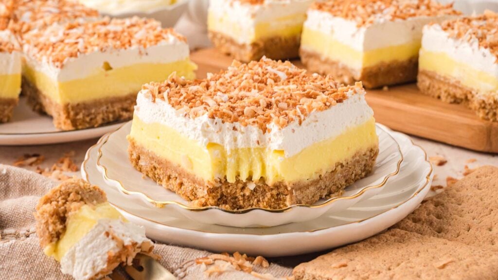 A square slice of layered coconut cream dessert bar with whipped topping and toasted coconut sits on a plate, with a forkful taken out. More dessert bars are in the background.