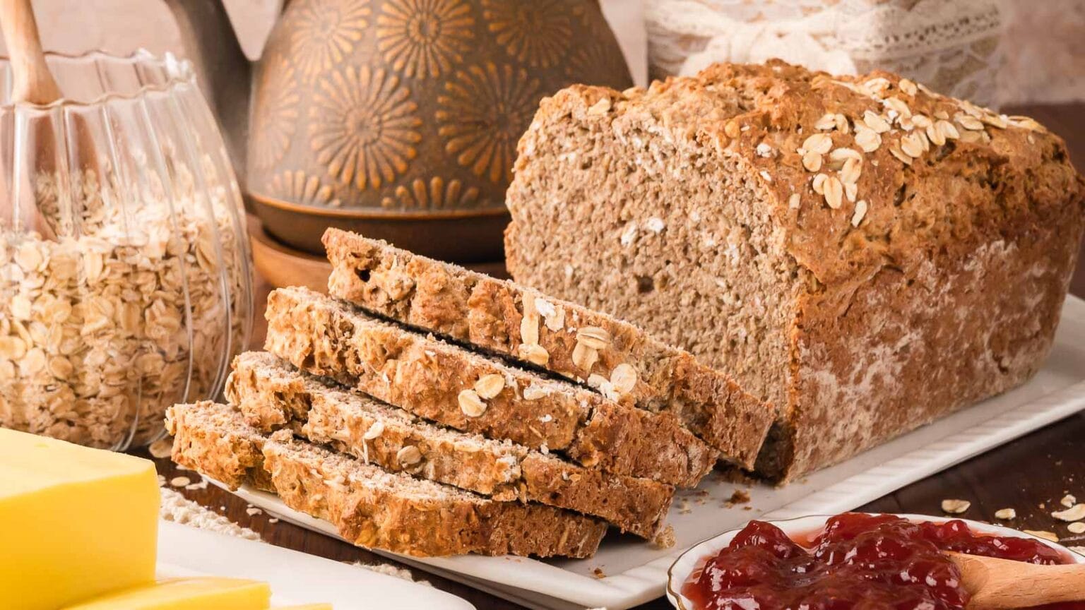 A loaf of oat bread with several slices cut sits on a white plate, surrounded by a bowl of oats, a block of butter, and a dish of red jam.