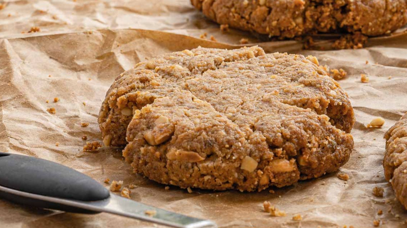 A large, chunky cookie with visible peanut pieces sits on crumpled brown parchment paper next to a metal spatula with a black handle.