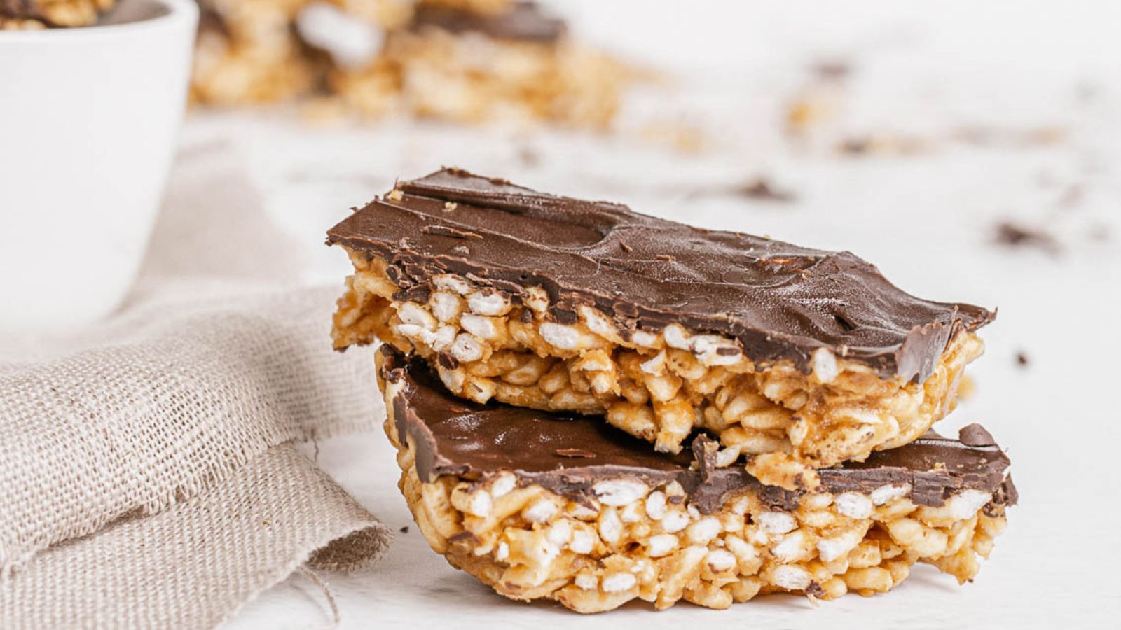 Two chocolate-covered puffed rice bars are stacked on a light surface with a piece of burlap cloth nearby.