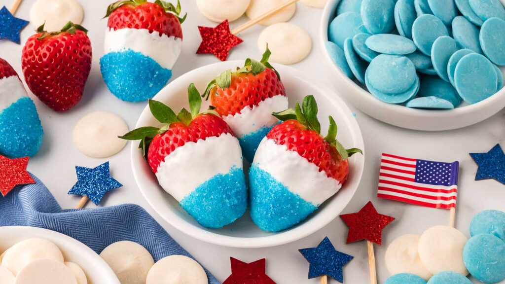 Strawberries dipped in white chocolate and blue sugar in a bowl, surrounded by blue candy melts, white candies, star decorations, and a small American flag.