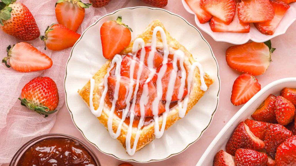 A strawberry danish with icing on a plate, surrounded by fresh strawberries and a bowl of sliced strawberries on a pink surface.