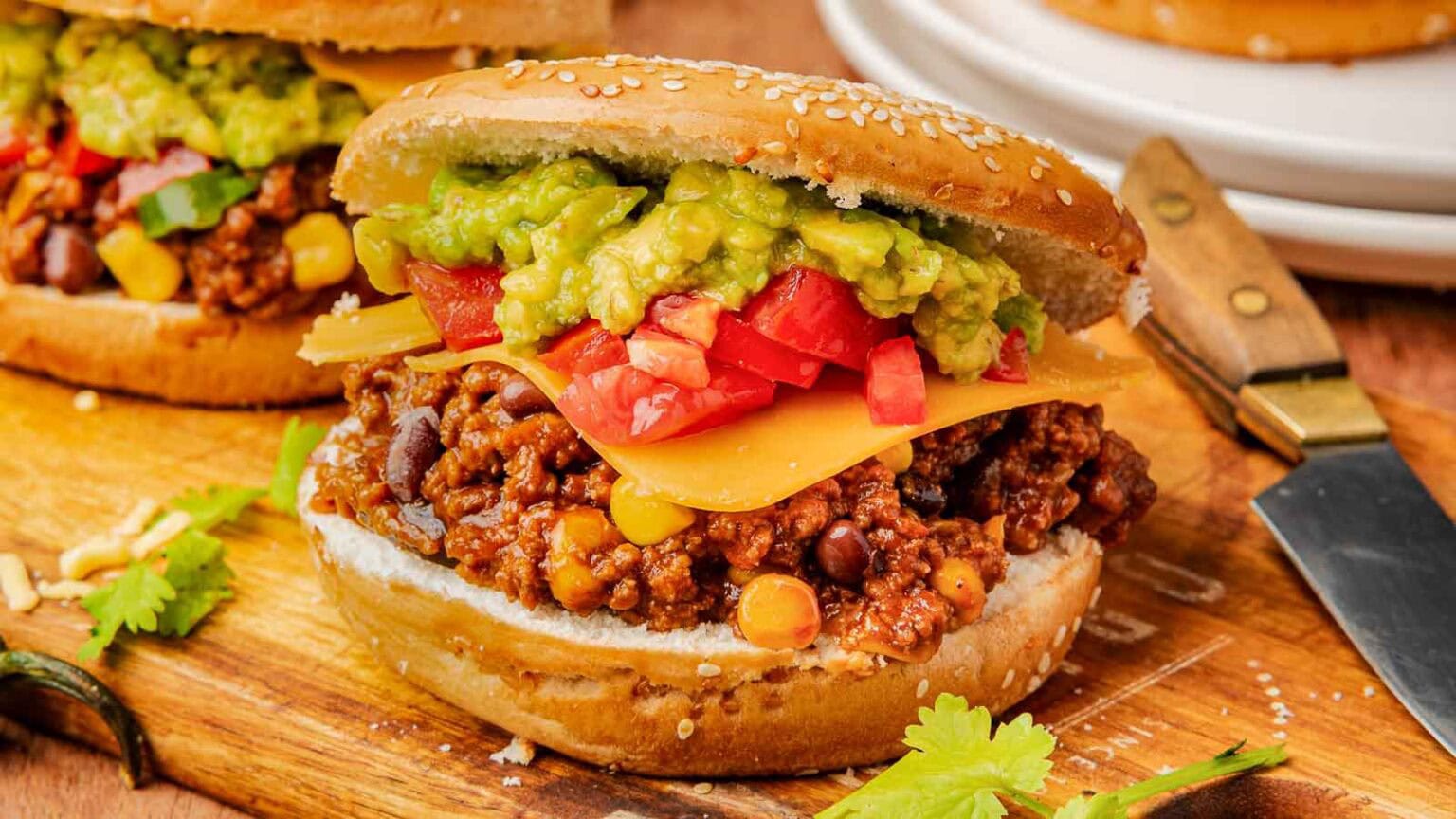 A sandwich bun filled with ground beef, black beans, corn, cheddar cheese, chopped tomatoes, and guacamole, placed on a wooden board next to a knife.