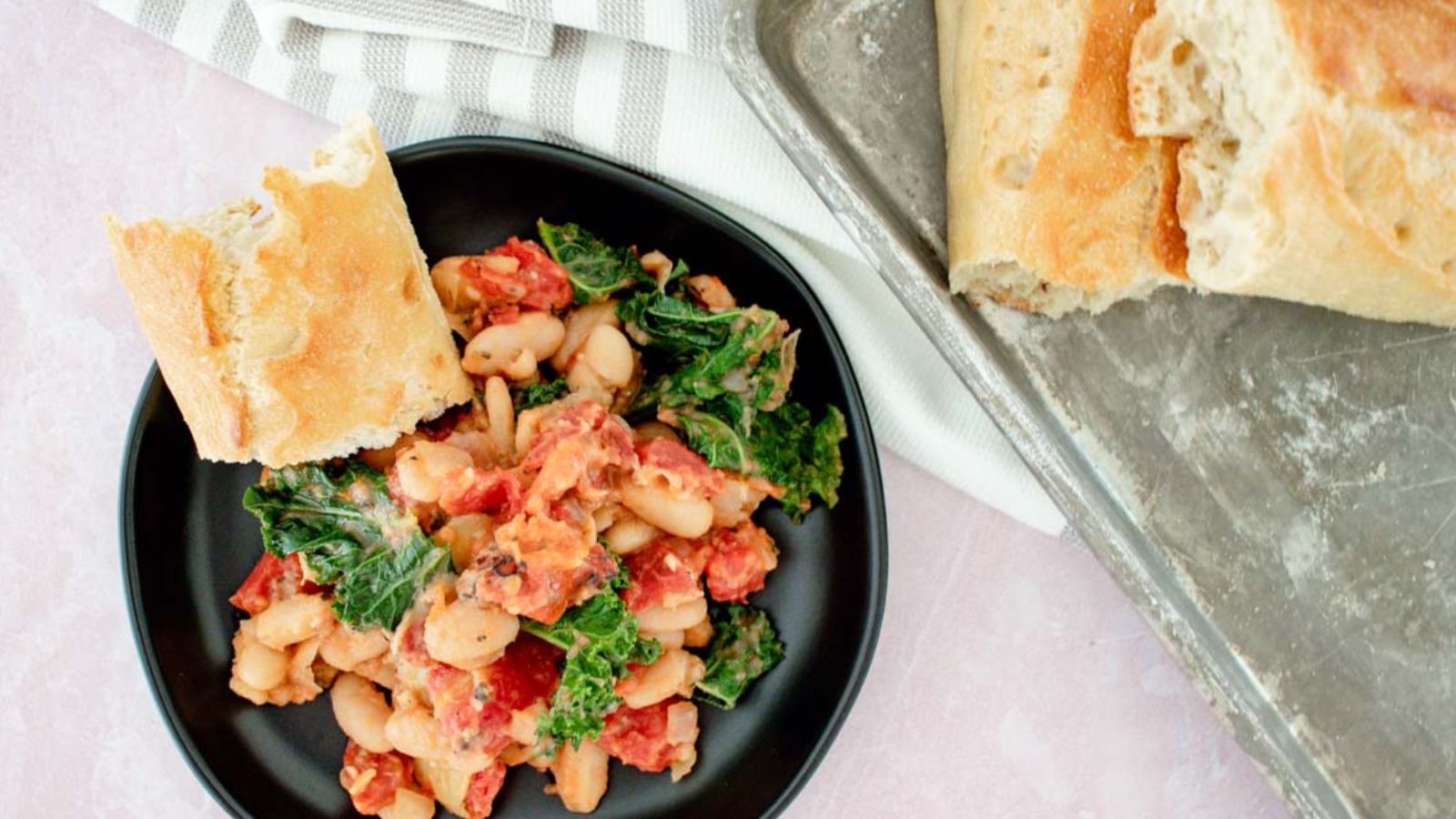 A black plate with a bean, tomato, and kale stew, served with a piece of crusty bread; additional bread is on a metal tray nearby.
