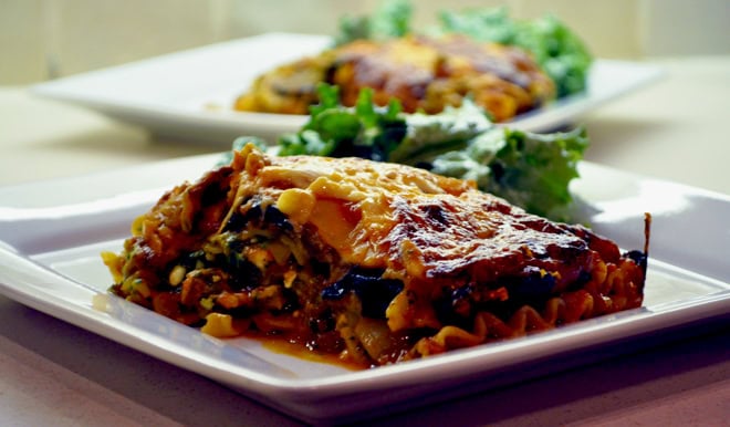 A slice of baked vegetable lasagna on a white plate, served with a side of leafy greens. Another plate with the same meal is blurred in the background.