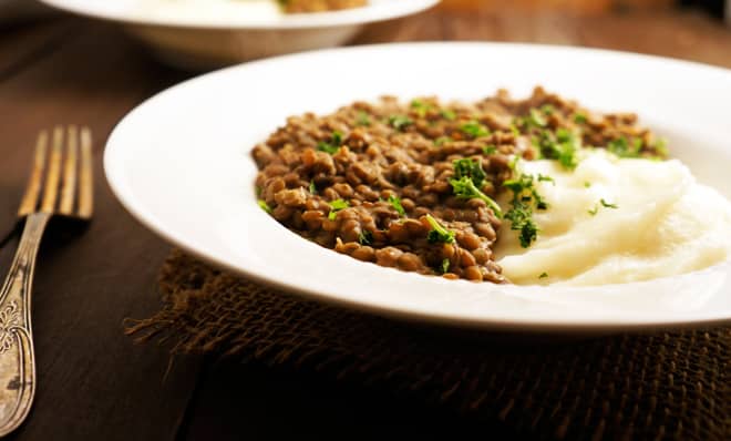 A white plate with mashed potatoes and a serving of cooked lentils garnished with chopped parsley, placed on a brown woven placemat.