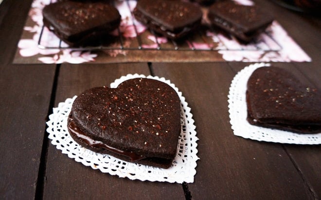 Heart-shaped chocolate cookies with chocolate filling are placed on paper doilies on a dark wooden surface, with more cookies cooling on a rack in the background.