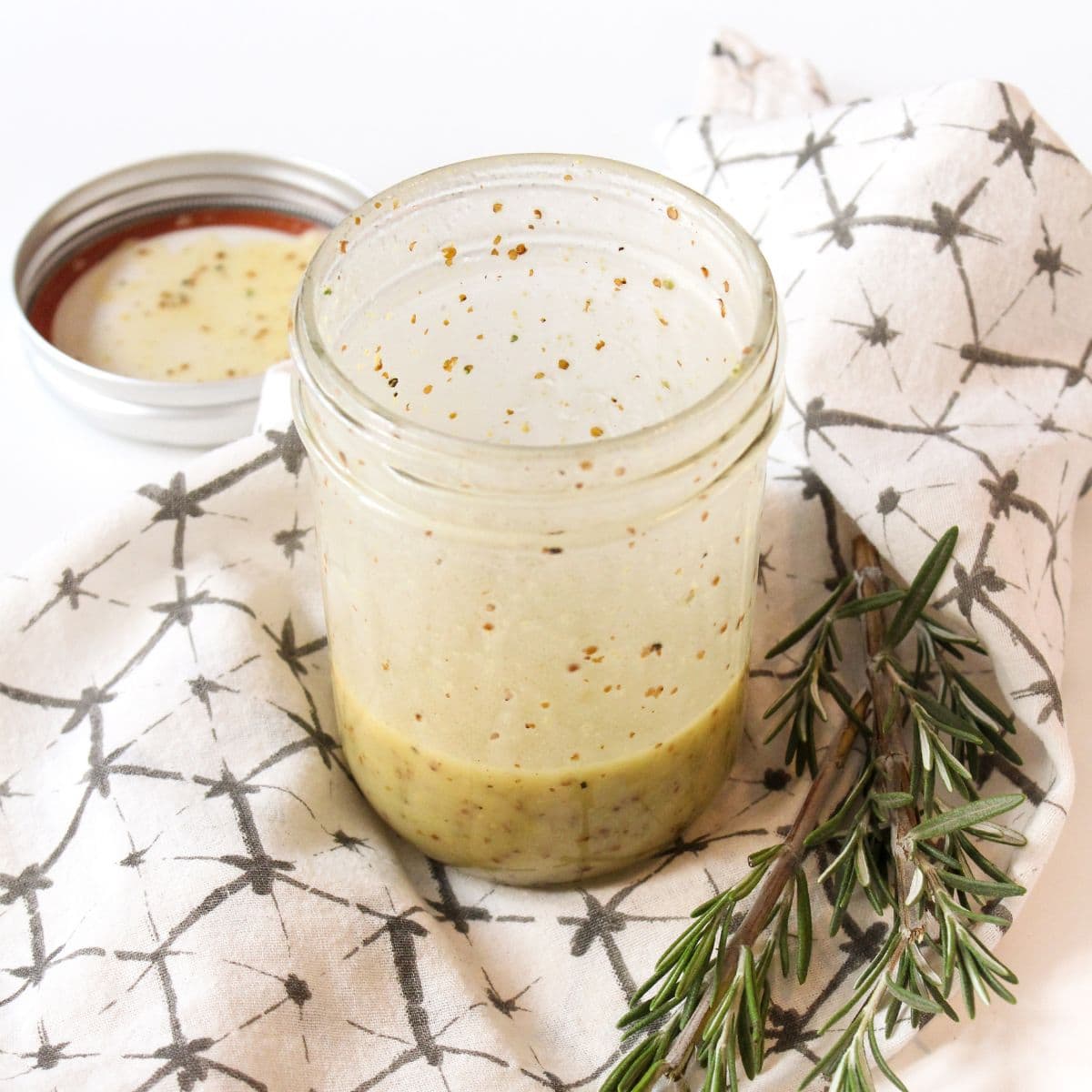 A glass jar with homemade salad dressing sits on a patterned cloth next to a sprig of rosemary and an open lid.