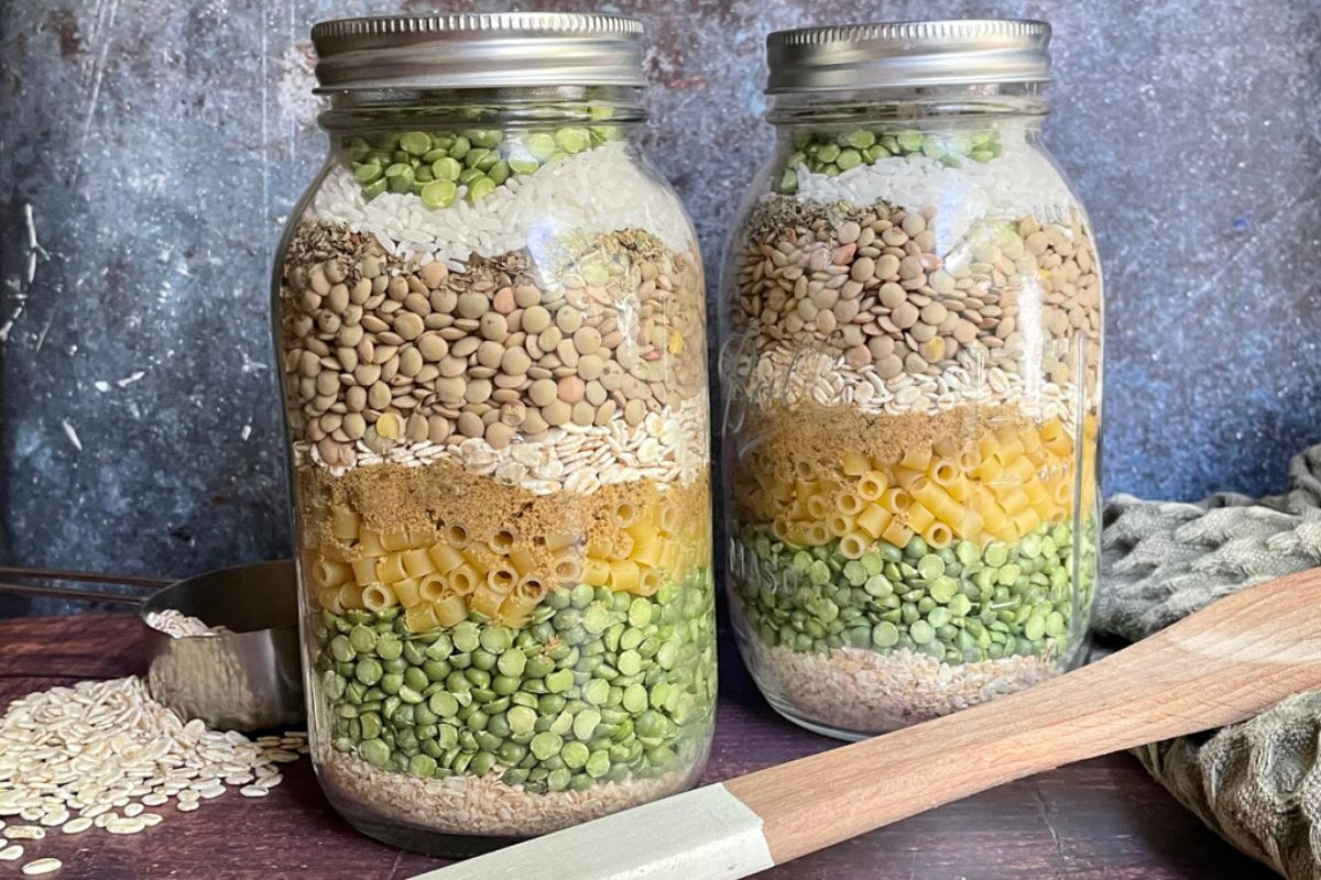 Two glass jars filled with layered dried ingredients, including split peas, pasta, lentils, rice, and oats, placed on a wooden surface with a scoop and cloth nearby.