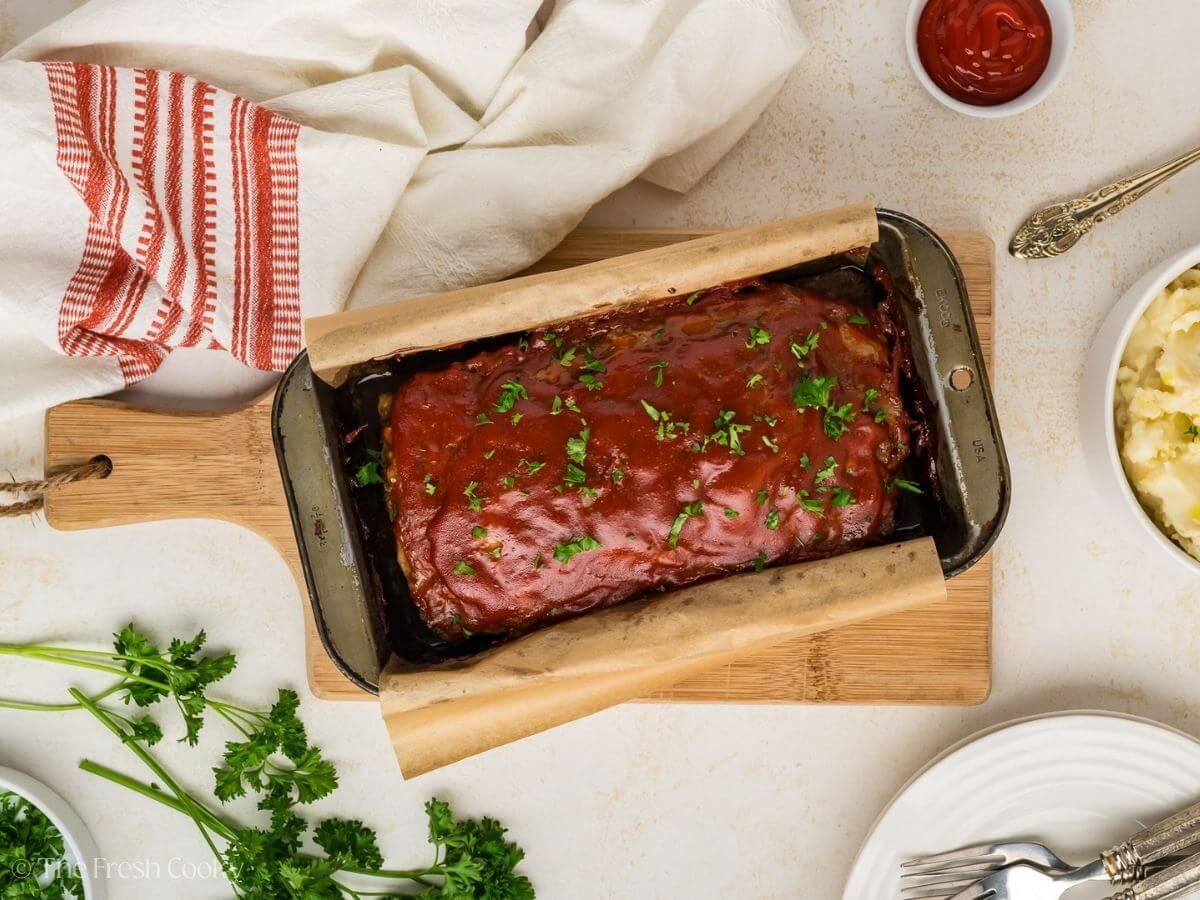 A glazed meatloaf in a parchment-lined loaf pan on a wooden board, garnished with chopped parsley, with mashed potatoes and a bowl of ketchup nearby.