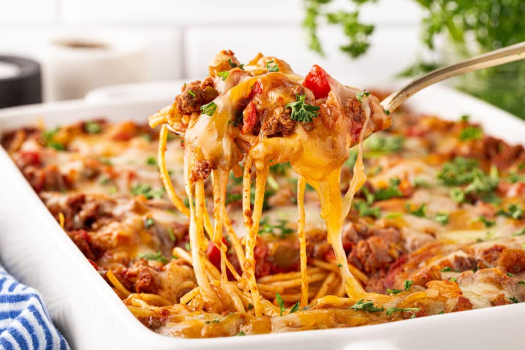 A fork lifts a serving of cheesy baked spaghetti with ground meat and tomato sauce from a white casserole dish, topped with melted cheese and chopped parsley.