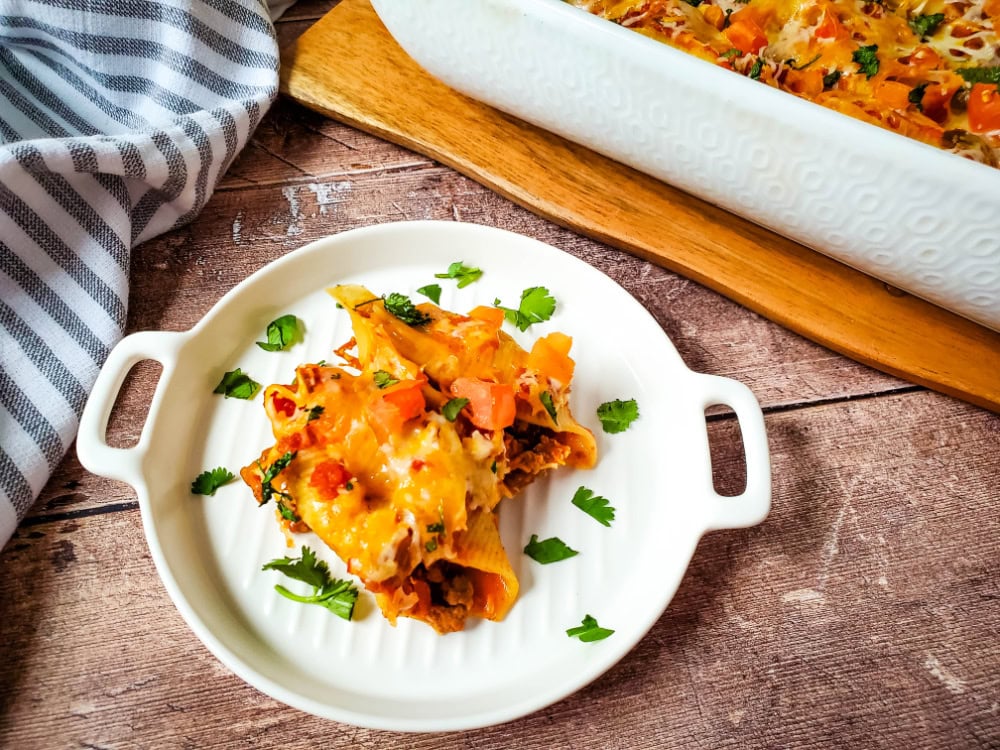 Stuffed pasta shells topped with melted cheese and diced vegetables on a white plate, garnished with herbs; casserole dish and striped towel in background.
