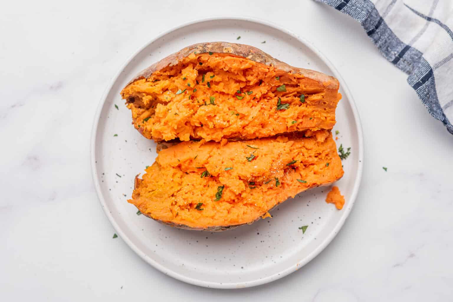 A baked sweet potato cut open and fluffed, sprinkled with chopped herbs, served on a white plate.