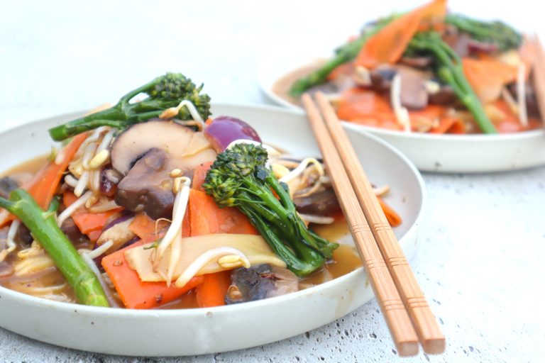 Two plates of stir-fried vegetables, including carrots, mushrooms, broccolini, and bean sprouts, served with wooden chopsticks on a light surface.