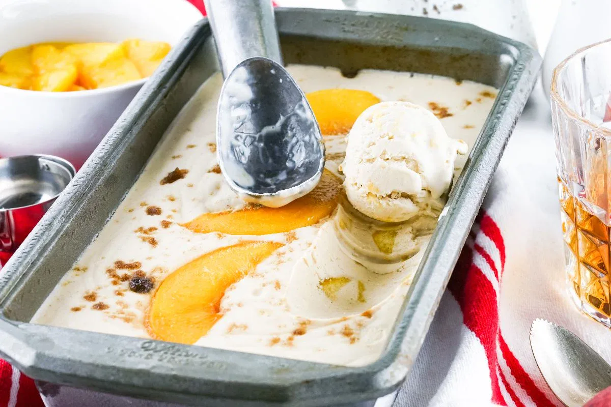 A metal pan filled with ice cream, peach slices, and a scoop, with a bowl of peaches and a glass of iced tea nearby.