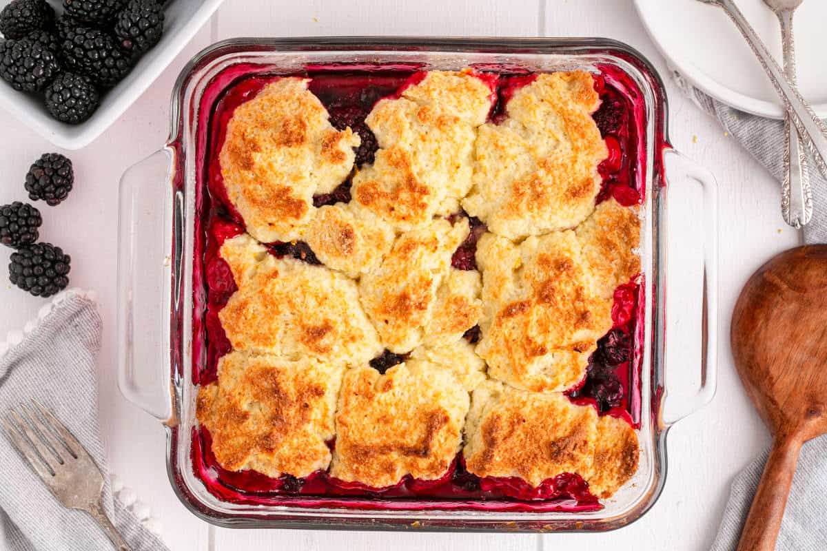 A square glass baking dish filled with blackberry cobbler, topped with golden-brown biscuit dough. Fresh blackberries, utensils, and a napkin are nearby on a white surface.