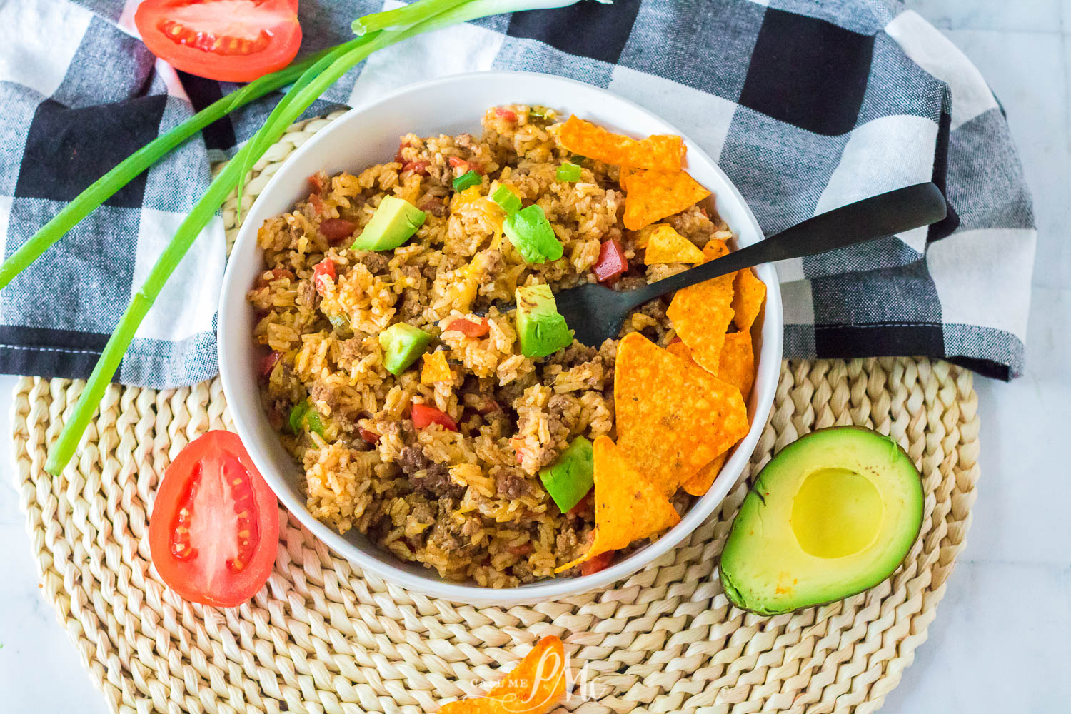 A bowl of seasoned rice with diced avocado, tomatoes, and tortilla chips on top, placed on a woven mat with a checkered cloth, green onions, and sliced avocado and tomato nearby.