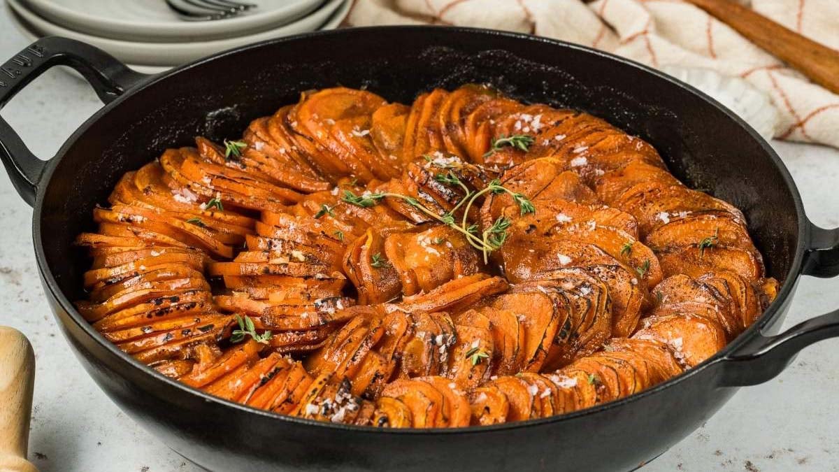 A cast iron skillet filled with neatly arranged, sliced roasted sweet potatoes garnished with herbs and sprinkled with coarse salt.