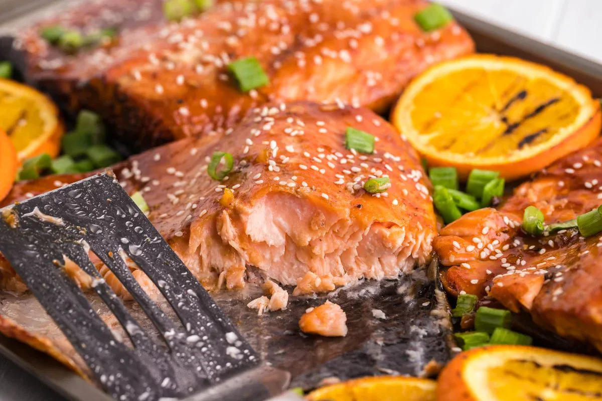 Baked salmon fillet with a glazed surface, garnished with sesame seeds, chopped green onions, and orange slices, partially served with a metal spatula on a tray.