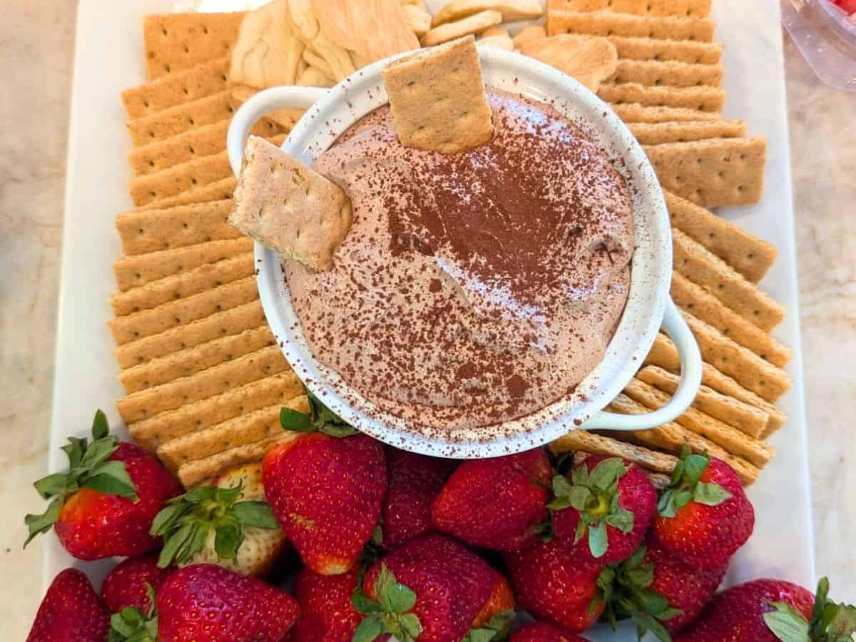 A bowl of chocolate dip sprinkled with cocoa powder, surrounded by graham crackers, apple slices, and fresh strawberries on a platter.